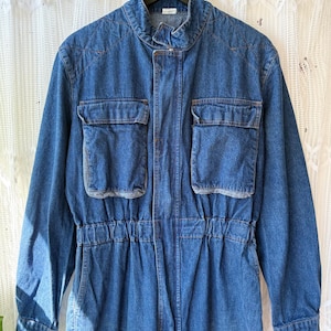 Könnte beinhalten: Blauer Jeans-Overall mit langen Ärmeln, Stehkragen und mehreren Taschen. Der Overall hat eine geraffte Taille und einen Frontreißverschluss. Das Kleidungsstück hängt an einem schwarzen Kleiderbügel.