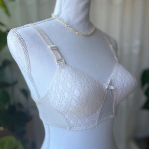 May include: Ivory-colored bra with a delicate lace pattern on the cups. The bra features adjustable straps and a small bow detail. A pearl necklace is draped around the mannequin's neck.