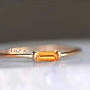 Può includere: Un delicato anello d'oro con una gemma arancione rettangolare. La gemma è incastonata orizzontalmente in una lunetta e la fascia è sottile e lucida. L'anello è esposto su una superficie bianca con uno sfondo sfocato.