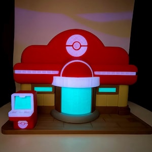 May include: A red and white building with a glowing blue center and a small red arcade machine. The building has a red roof with a white circle and a white stripe. The arcade machine is red with a white screen.