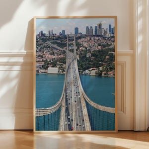 May include: A framed print of a suspension bridge over a body of water, with a city skyline in the background. The bridge is filled with cars and trucks. The water is a deep blue, and the sky is a light blue with some clouds.