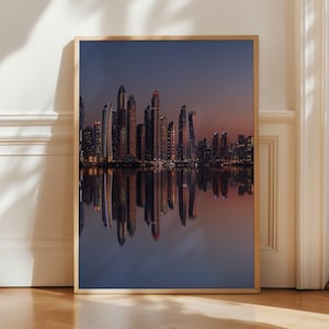 May include: A framed print of a cityscape at dusk. The image features a skyline of tall buildings with illuminated windows reflected in the water below. The sky transitions from a deep blue to a warm orange hue.