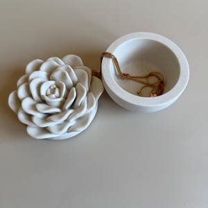 Peut inclure: Un vide-poches décoratif blanc avec une base en forme de rose et un bol rond. Un collier et des boucles d'oreilles dorés reposent à l'intérieur du bol. Le plat est fait d'un matériau lisse et mat.