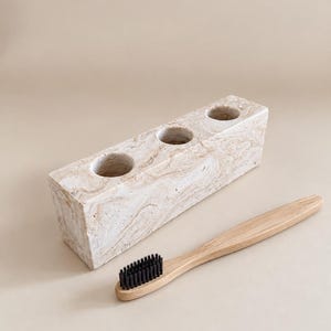 Puede incluir: Un soporte rectangular de piedra veteada en tonos beige y crema con tres aberturas circulares alineadas, acompañado de un cepillo de dientes con mango de madera marrón claro y cerdas negras situado frente a la base de piedra.