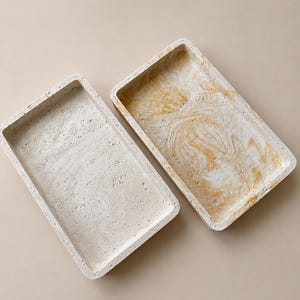 Könnte beinhalten: Zwei rechteckige Tabletts mit poröser, steinartiger Textur. Ein Tablett ist in einem einfarbigen Off-White gehalten, während das andere ein marmoriertes Muster mit Wirbeln in Weiß und Goldgelb aufweist, beide mit erhöhtem Rand und flachem Boden.