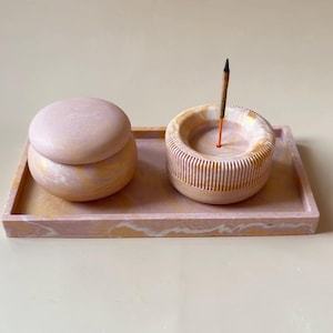 Op de afbeelding: Een rechthoekig dienblad met een gemarmerde roze, witte en gouden afwerking met een ronde container met deksel en een ronde wierookhouder met verticaal geribbelde zijkanten. Een dun wierookstokje staat in het midden van de houder.