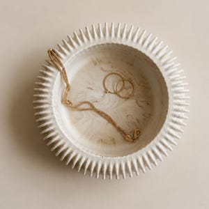Op de afbeelding: Een ronde, gebroken witte keramische kom met een getextureerde, stekelige rand en een gemarmerde binnenkant. In de kom liggen een delicate gouden ketting en twee dunne gouden ringen, wat een eenvoudig arrangement van sieraden in de schaal vormt.