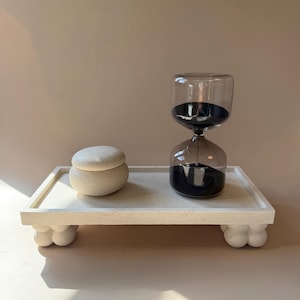 Puede incluir: Una bandeja rectangular de color blanco roto con motas y cuatro patas redondeadas en forma de burbuja. Sobre la bandeja hay un pequeño tarro con tapa a juego y un reloj de arena de cristal con arena negra y marco de cristal gris oscuro.