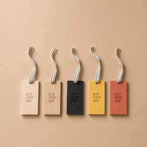 Può includere: Cinque etichette rettangolari color crema, beige, nero, giallo e terracotta, ciascuna con il testo in rilievo "err eke rre" e un nastro a righe bianche e nere fissato in alto per appenderle, disposte in una fila orizzontale.