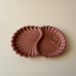 Puede incluir: Una bandeja de color terracota con dos compartimentos en forma de dos conchas de vieira unidas. La bandeja presenta una superficie texturizada y acanalada que irradia desde el centro de cada sección. Dos anillos finos de tono dorado descansan en su interior.