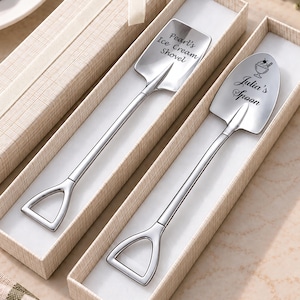 Puede incluir: Dos cucharas de metal plateado con forma de palas en miniatura, cada una en una caja de regalo beige texturizada. Una cuchara está grabada con "Pearl's Ice Cream Shovel" y la otra con "Julia's Spoon" junto a un icono de una copa de helado.