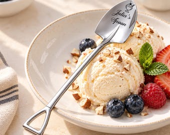 May include: A silver-toned metal spoon shaped like a miniature shovel, featuring an engraved graphic of a sundae and the text "Julia's Spoon" on the bowl. The handle ends in a triangular grip, resting on a bowl of vanilla ice cream with berries.