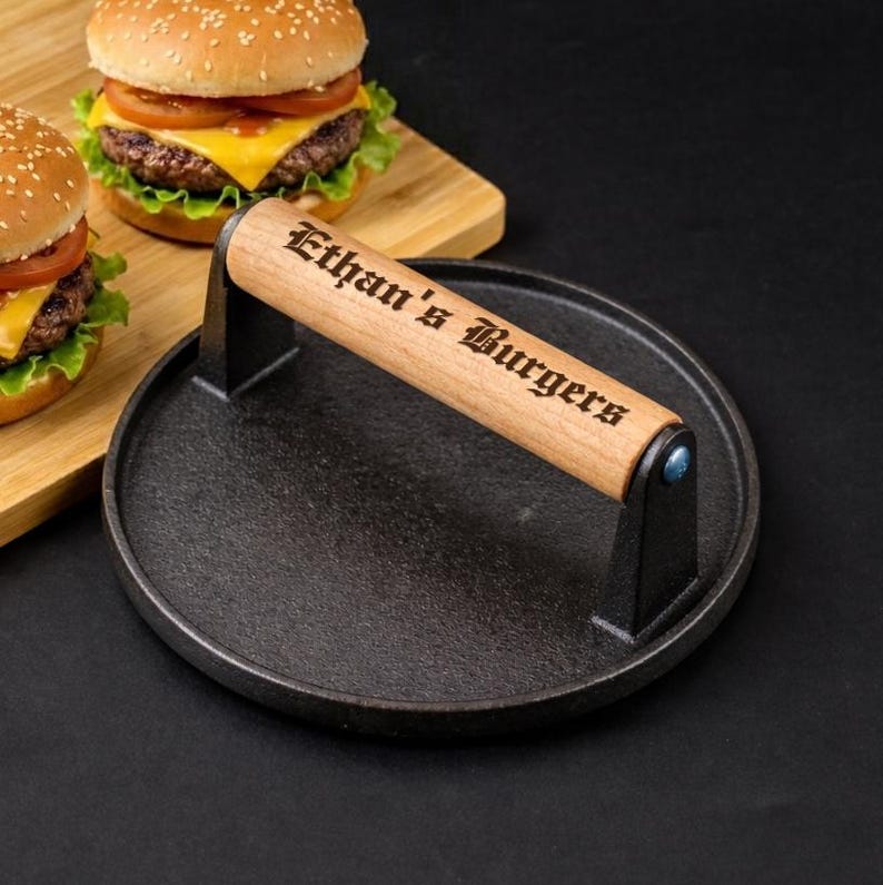 Pode incluir: Uma prensa para hamb&uacute;rgueres em ferro fundido preto com uma pega de madeira gravada com "Ethan's Burgers". A prensa &eacute; redonda e tem uma pega presa com suportes de metal preto. Dois hamb&uacute;rgueres est&atilde;o no fundo numa t&aacute;bua de cortar de madeira.