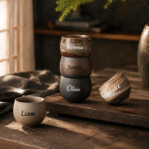 May include: Five rounded ceramic cups with textured finishes in shades of brown, grey, and beige. Each cup is personalised with a name in a distinct font: Emma, James, Olivia, Blake, and Liam. The cups are arranged in a stack and individually.