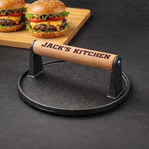 Pode incluir: Uma prensa para hamb&uacute;rgueres em ferro fundido preto com uma pega de madeira gravada com "JACK'S KITCHEN". Dois cheeseb&uacute;rgueres com p&atilde;es de s&eacute;samo, alface, tomate e queijo est&atilde;o numa t&aacute;bua de madeira no fundo. A prensa &eacute; redonda.