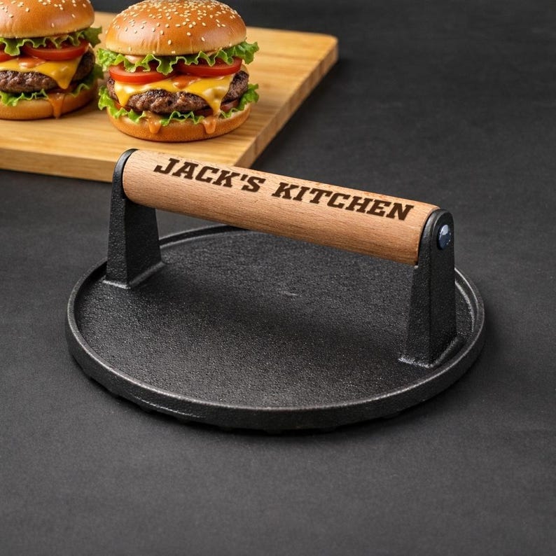 Pode incluir: Uma prensa para hamb&uacute;rgueres em ferro fundido preto com uma pega de madeira gravada com "JACK'S KITCHEN". Dois cheeseb&uacute;rgueres com p&atilde;es de s&eacute;samo, alface, tomate e queijo est&atilde;o numa t&aacute;bua de madeira no fundo. A prensa &eacute; redonda.