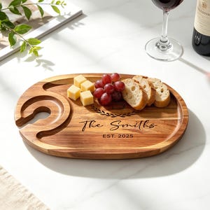 Könnte beinhalten: Ovales Servierbrett aus Holz mit Weinglashalter. Das Brett ist mit "The Smiths" und "EST. 2025" graviert. Es enthält Käsewürfel, rote Trauben und geschnittenes Brot. Ein Weinglas und eine Flasche befinden sich im Hintergrund.