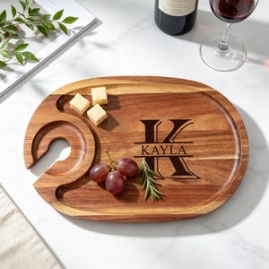 Op de afbeelding: Een ovale houten serveerschaal met een wijnglashouder en de naam "Kayla" onder een grote initiaal "K". De schaal bevat kaas, druiven en rozemarijn. Het hout heeft een natuurlijke nerf.