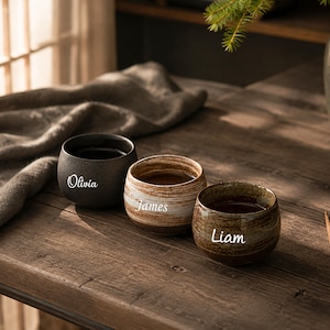May include: Three rounded ceramic cups with a smooth finish. The first cup is matte black with the name Olivia in white script. The second cup features a white and brown marbled pattern with the name James. The third cup is brown with the name Liam.