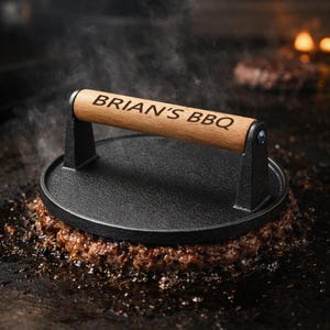 May include: A black cast iron burger press with a light brown wooden handle, engraved with "BRIAN'S BBQ", is pressing a burger patty. Steam rises from the cooking meat. The press is designed for grilling burgers.