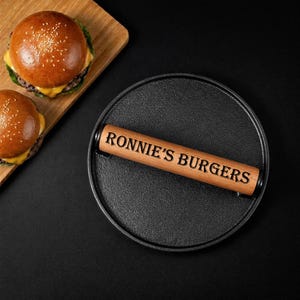 Pode incluir: Uma prensa para hamb&uacute;rgueres de ferro fundido com uma pega de madeira gravada com "RONNIE'S BURGERS" repousa sobre uma superf&iacute;cie preta. Dois hamb&uacute;rgueres numa t&aacute;bua de cortar de madeira est&atilde;o no fundo. Os hamb&uacute;rgueres t&ecirc;m p&atilde;es de s&eacute;samo e queijo.