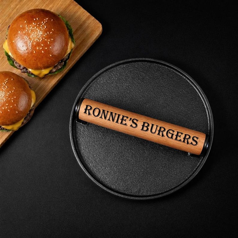 Pode incluir: Uma prensa para hamb&uacute;rgueres de ferro fundido com uma pega de madeira gravada com "RONNIE'S BURGERS" repousa sobre uma superf&iacute;cie preta. Dois hamb&uacute;rgueres numa t&aacute;bua de cortar de madeira est&atilde;o no fundo. Os hamb&uacute;rgueres t&ecirc;m p&atilde;es de s&eacute;samo e queijo.
