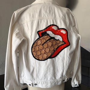 May include: A white denim jacket with a large, sequined patch on the back depicting red lips and a tongue with a brown pattern. The jacket has a classic design with button closures and pockets.