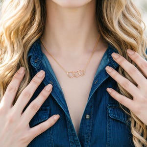 Puede incluir: Un collar dorado con tres colgantes de corazón entrelazados, cada uno adornado con pequeños detalles brillantes. El collar se muestra sobre una camisa vaquera. Una joya delicada.