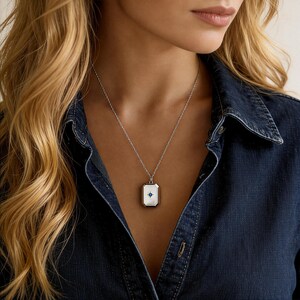 May include: Silver necklace with a rectangular pendant. The pendant has a white background with a blue starburst design in the center. The necklace is worn on a person wearing a denim shirt.