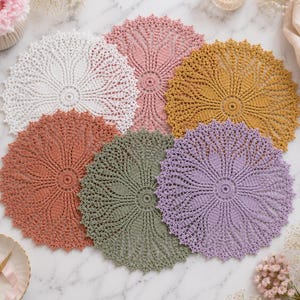 May include: A collection of six circular crochet doilies in white, dusty pink, mustard yellow, terracotta, sage green, and lavender. Each doily features a detailed, intricate floral-inspired pattern with a central medallion and scalloped edges.