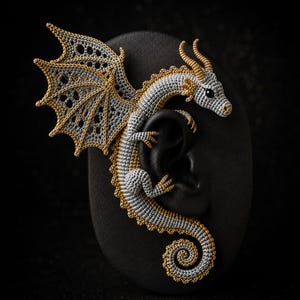 May include: A dragon-shaped ear cuff crafted from silver and gold thread. The creature features a detailed head with horns, a large wing with intricate openwork, and a long, curled tail. The body is textured with fine stitching throughout.