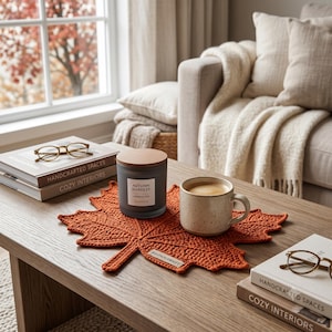 May include: A dark grey candle with a wooden lid labelled Autumn Harvest Amber & Oak, a speckled beige ceramic mug, and a pair of gold-rimmed glasses resting on a stack of books, all arranged on a burnt orange crocheted maple leaf coaster.