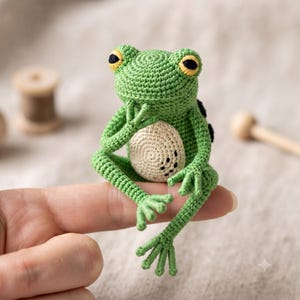 May include: A small, green, crocheted frog figurine with a cream-colored belly, yellow and black eyes, and long, slender limbs. The frog is posed in a seated position with one hand near its chin, showcasing intricate yarn stitching throughout.