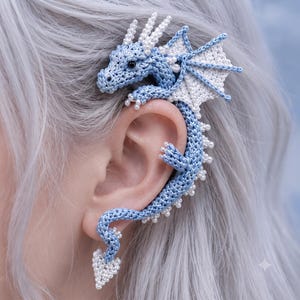 May include: A crochet ear cuff shaped like a dragon, featuring a light blue body with white wings, white beaded accents along the spine, and a white beaded tail tip. The dragon is positioned to wrap around the outer edge of the ear structure.
