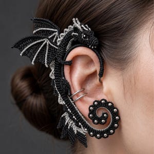 May include: A black and silver crocheted dragon ear cuff that wraps around the outer ear. The dragon features detailed wings, a textured body, and a coiled tail adorned with small silver beads. Two plain silver rings are visible on the earlobe.