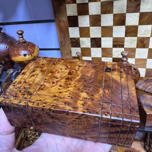 Könnte beinhalten: Eine handgefertigte Holzkiste mit einer reichen, warm getönten Maserung. Die Kiste hat eine rechteckige Form mit Deckel und ist mit helleren Holzeinlagen versehen. Im Hintergrund ist ein Schachbrett aus Holz zu sehen.