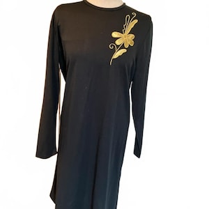 May include: Black long-sleeve dress with a gold floral design on the chest. The dress has a round neckline and a slightly asymmetrical hemline. The gold embroidery adds a touch of elegance to the simple, classic design.