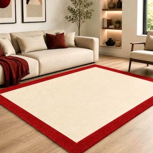 May include: A rectangular area rug featuring a large, solid cream-colored center panel framed by a wide, vibrant red border. The rug has a flat-woven texture and is positioned on a light-toned wooden floor, providing a clean, geometric contrast.
