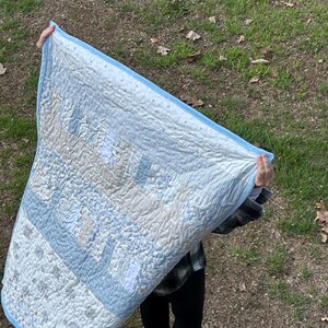 May include: A light blue and white patchwork quilt with a blue border. The quilt features various patterns, including stripes and small animal prints. The quilt is held up, showcasing its design and texture.