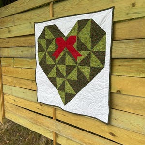 May include: A square quilt featuring a large heart shape constructed from dark green and light green triangular fabric patches. A bright red fabric bow is appliqued in the center of the heart. The heart is set against a white background.