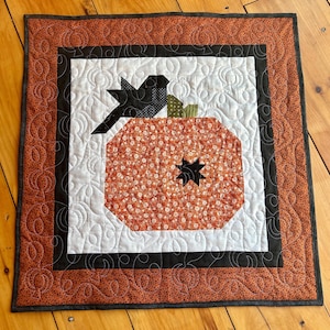 May include: A square quilted wall hanging featuring a central orange pumpkin with a small black star accent, a black stem, and a green leaf. The pumpkin is set against a white background with black swirl quilting and a wide orange border.