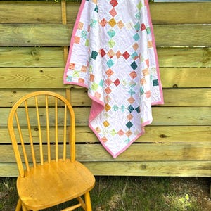 May include: A handmade patchwork quilt featuring a light lavender background with a repeating diamond pattern composed of small squares in various colors including teal, orange, yellow, and green, finished with a solid pink fabric border.