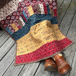 May include: A patchwork quilt with a Western theme, featuring brown, red, teal, and gold fabrics. The quilt has various patterns, including cowboy imagery and geometric designs. Brown leather cowboy boots are placed at the bottom of the quilt.