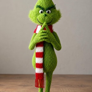 May include: A handmade green crochet figure of a character with a mischievous grin, wearing a red and white striped scarf. The figure has a pointed head, green eyes, and is posed with hands clasped. The figure is standing on a wooden surface.