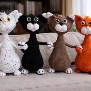 May include: Four handmade crochet cat dolls in white, black, brown, and orange. Each cat has large eyes, a white muzzle, and a long body. The cats are holding hands and are sitting on a beige couch. The dolls are a whimsical home decor item.