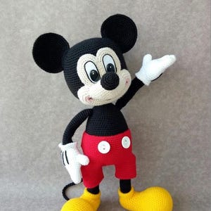 May include: Crocheted Mickey Mouse doll with black head and ears, a white face, and a black body. The doll wears red shorts with white buttons, white gloves, and yellow shoes. The doll is handmade and stands approximately 12 inches tall.