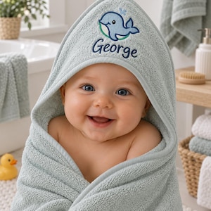 May include: A sage green hooded towel featuring an embroidered blue whale graphic and the name George in dark blue script. The towel is made of a soft, textured terry cloth fabric with a finished edge along the hood and body for a clean look.