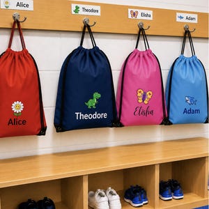 May include: Four drawstring bags in red, navy blue, pink, and light blue, each featuring a unique graphic and a name. The red bag has a flower, the navy bag a dinosaur, the pink bag a butterfly, and the light blue bag a shark graphic.