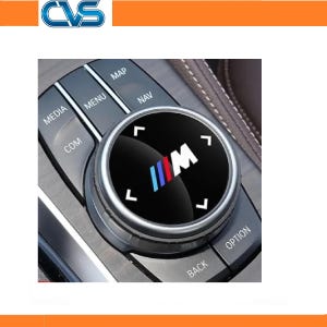 May include: A black circular car multimedia control knob featuring the M logo with blue, purple, and red stripes. The knob is surrounded by buttons labelled MEDIA, MENU, NAV, COM, BACK, and OPTION, with white directional arrows on the knob surface.