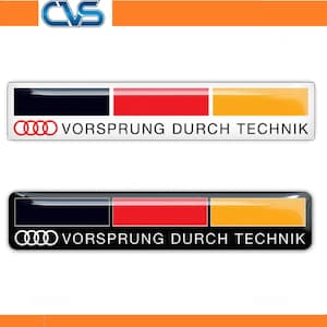 May include: Two rectangular car emblems featuring the Audi logo and the text VORSPRUNG DURCH TECHNIK. The top emblem has a white background with black, red, and yellow stripes. The bottom emblem has a black background with the same coloured stripes.